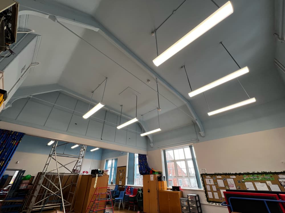 Newly installed LED pendant lighting suspended from the vaulted ceiling of a hall at Brigg Infant School, with a scaffold tower still in place during the works.