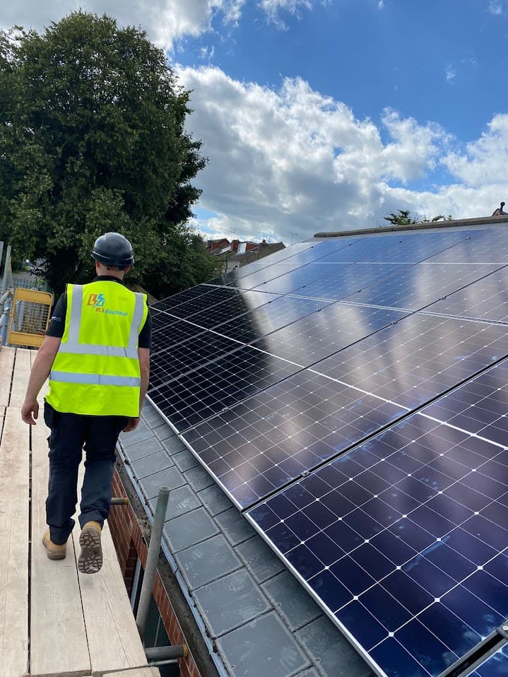 An RS Electrical engineer in high-visibility workwear and a hard hat walking along a roof access platform beside a newly installed photovoltaic array at Brigg Infant School, with trees and a partly cloudy sky in the background.