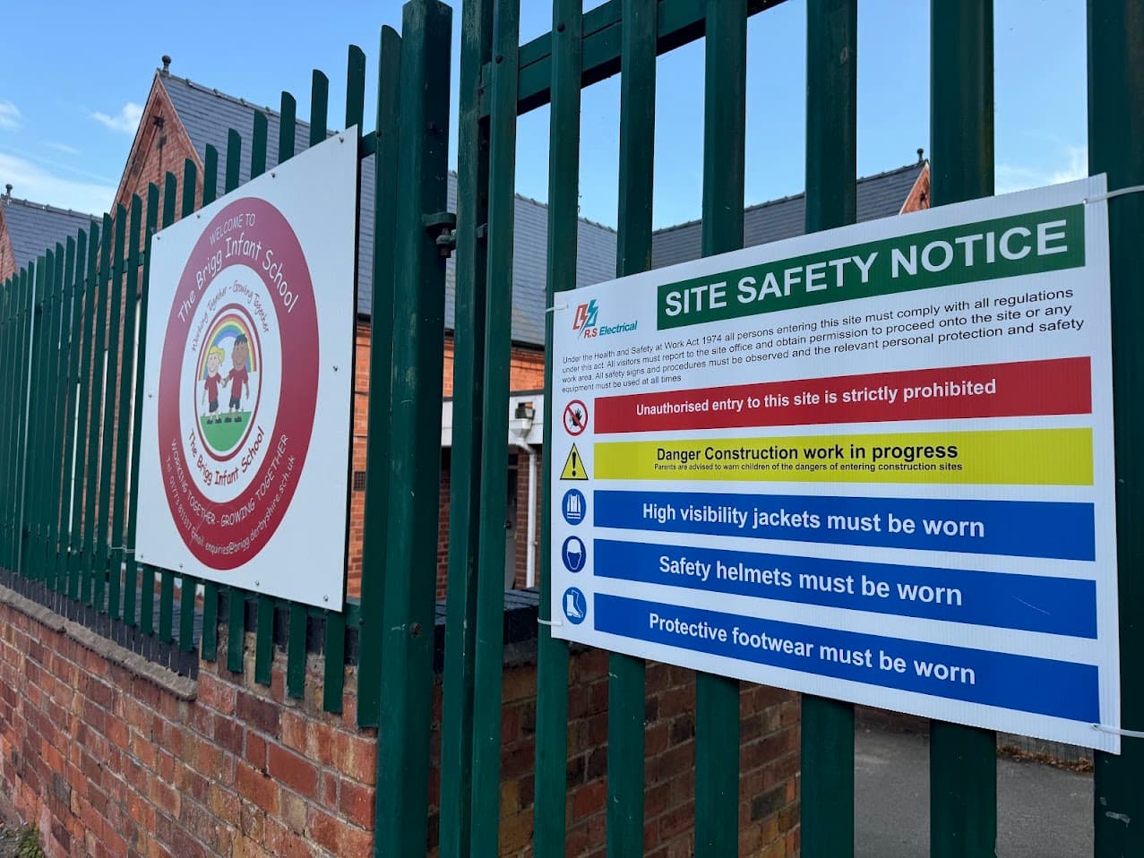 Site entrance at Brigg Infant School showing the school's welcome sign alongside an RS Electrical site safety notice attached to the perimeter fencing.