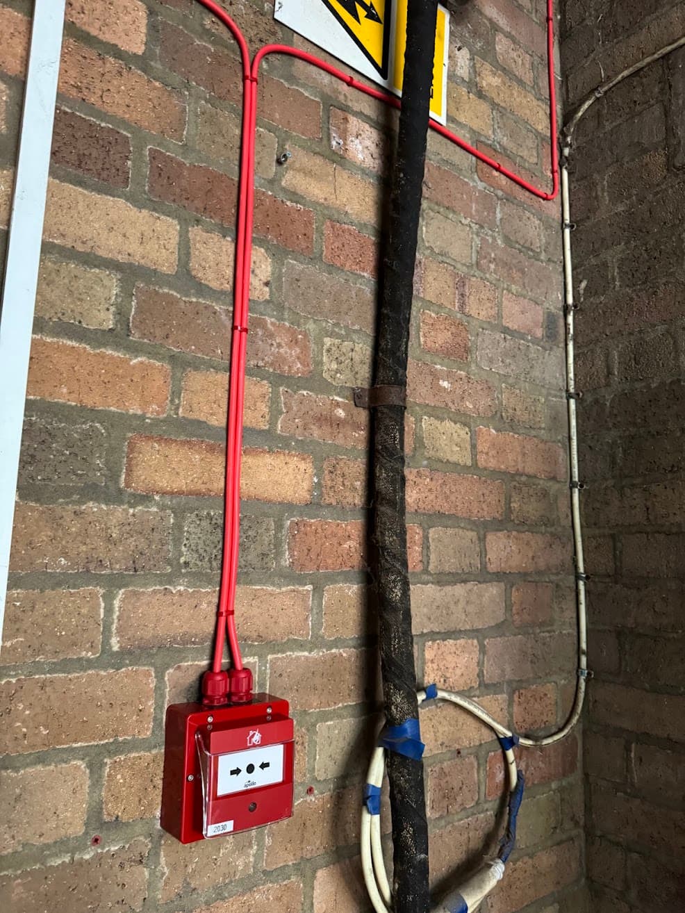 Fire alarm cabling and a wall-mounted Apollo manual call point installed against an exposed brick wall at Erdington Fire Station, surface-routed alongside existing pipework.