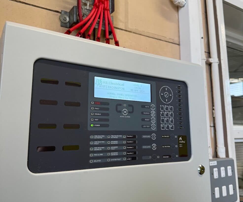 Wall-mounted addressable fire alarm control panel at Erdington Fire Station, with the display reading 'RS Electrical, WMFS Erdington, Normal Panel Operation' and fire alarm cabling entering above.