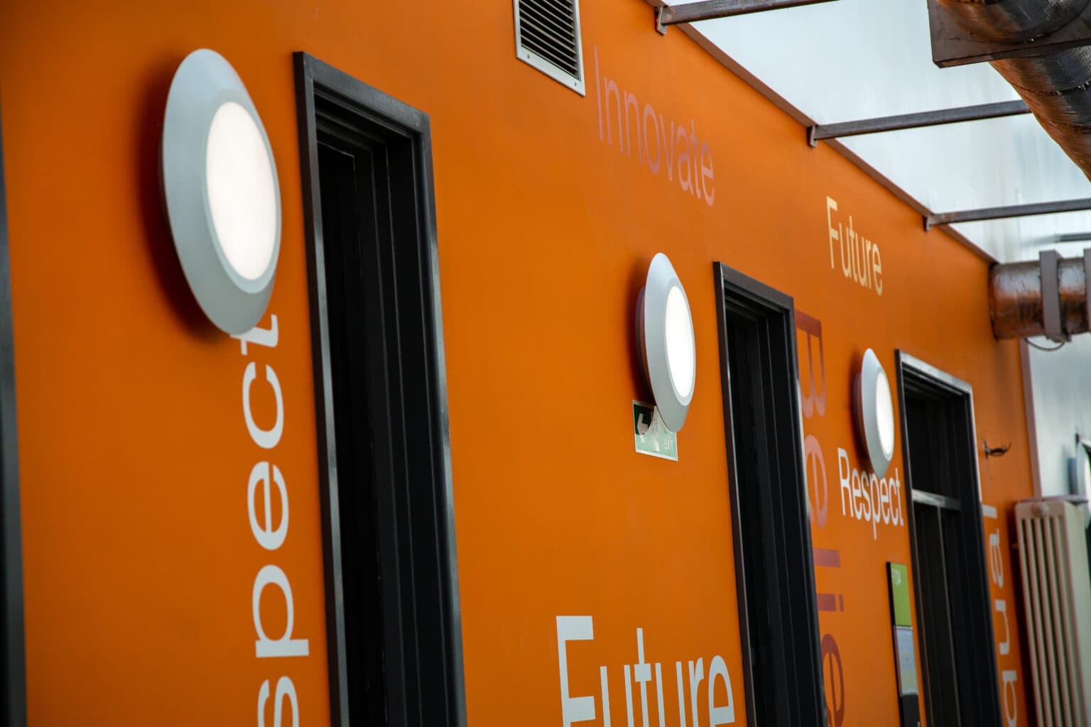Close-up of circular LED wall-mounted luminaires installed above doorways on an orange feature wall at The Foster Centre.