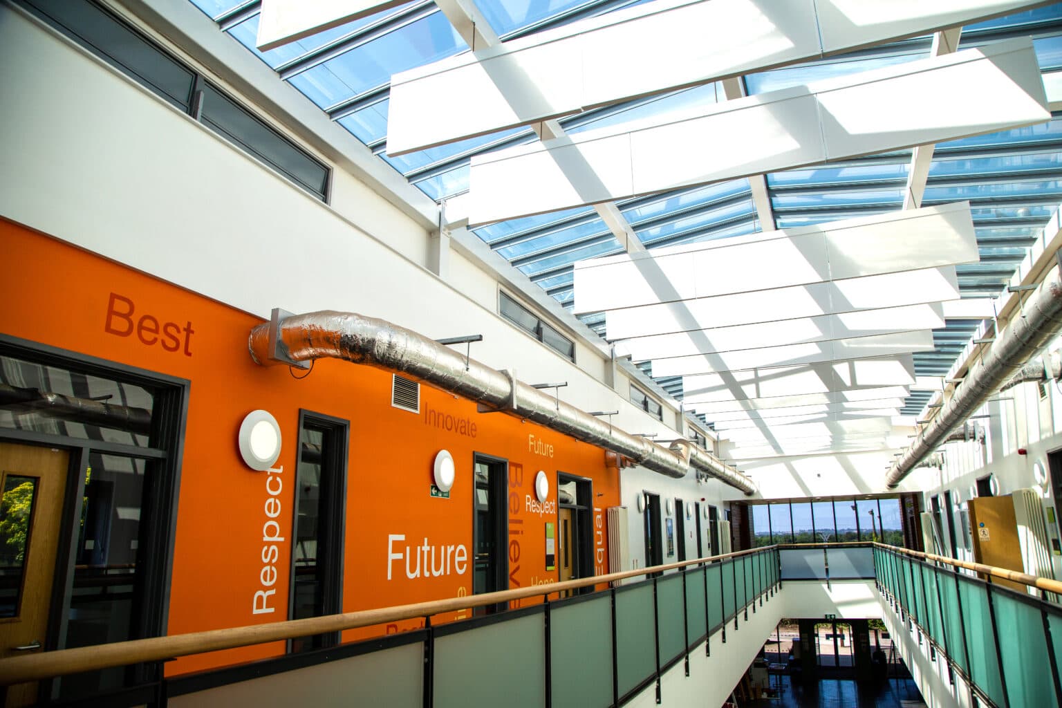 Interior view of a glazed circulation walkway at The Foster Centre, showing new circular LED wall luminaires alongside an orange feature wall and exposed ductwork beneath a glazed roof.