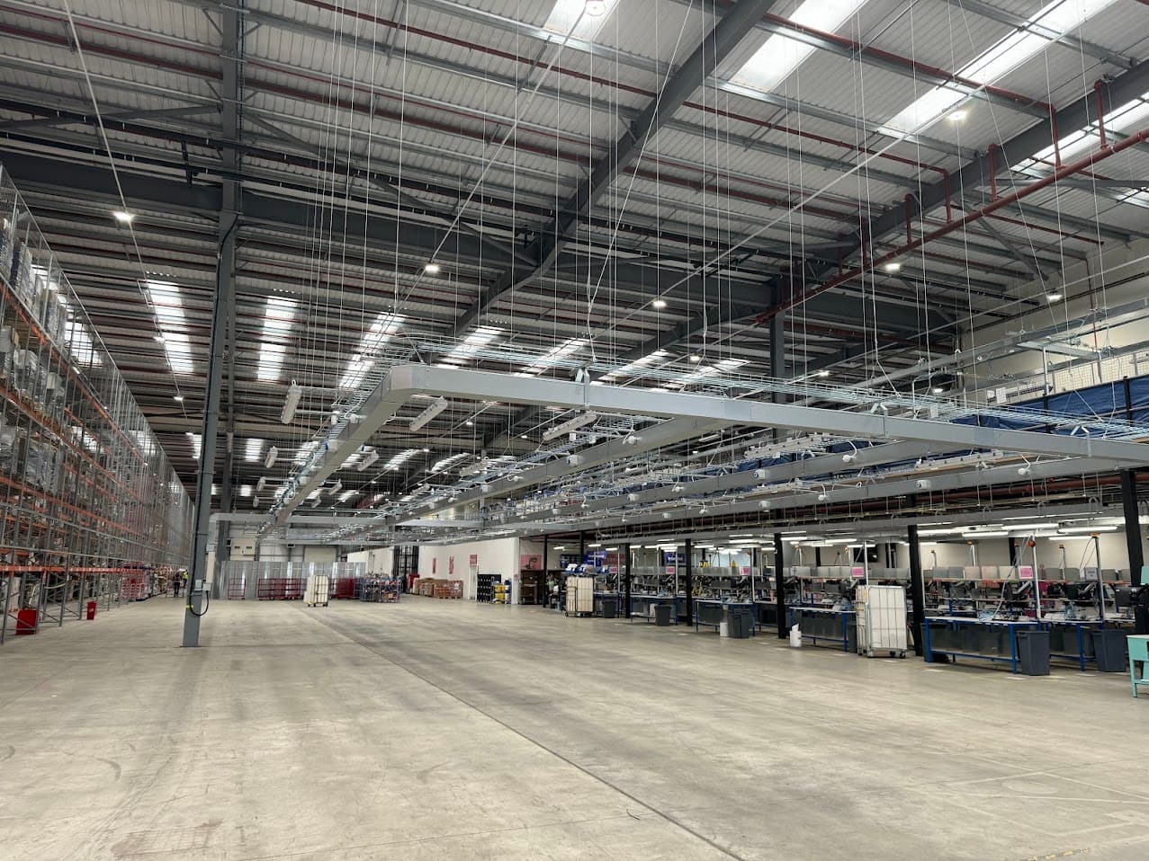 Wide view of the completed electrical containment, power and data infrastructure above the packing bench area at Moonpig's Tamworth distribution facility, with pallet racking to the left, packing bench workstations to the right, and a member of the site team visible in the aisle.