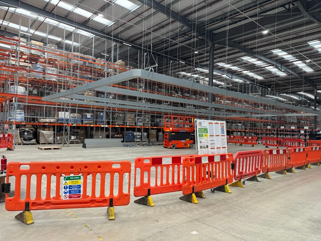 Overhead electrical containment installation above an active area of Moonpig's Tamworth warehouse, with temporary safety barriers, a scissor lift and site safety signage in the foreground, reflecting work carried out around the live operation.