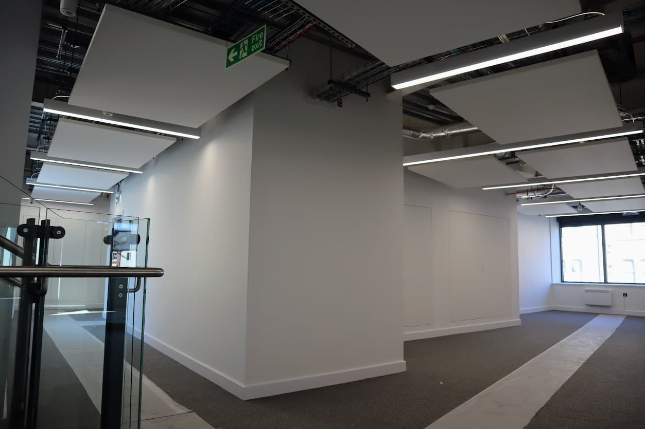 Completed lighting installation above an open ceiling void at Nuneaton Digital Skills Centre, with suspended acoustic rafts, linear LED luminaires and a glazed stair balustrade.