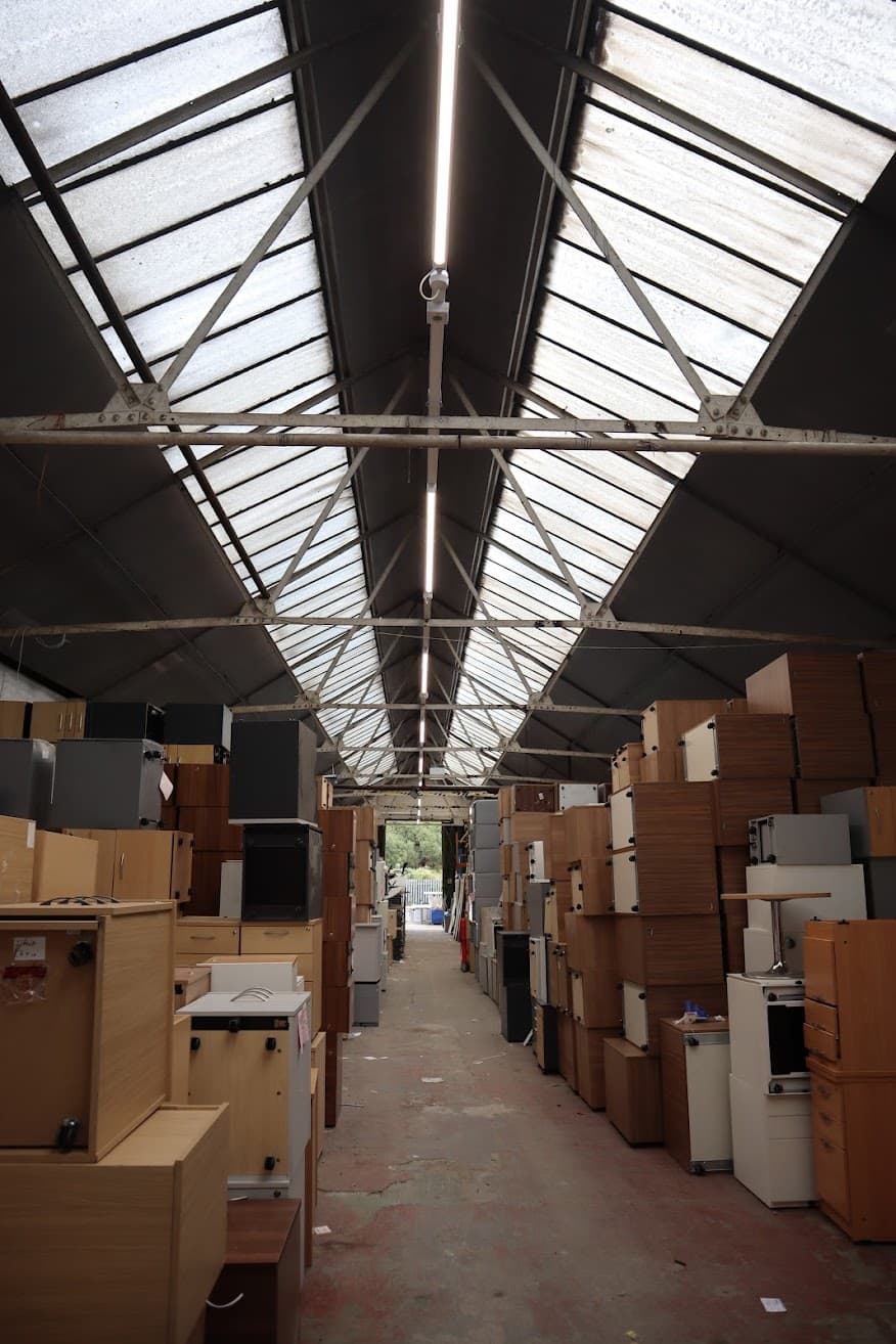 Wide view along the central aisle of ORS's warehouse in Burton-on-Trent, showing new linear LED lighting installed along the roof apex above stacked office furniture stored either side.