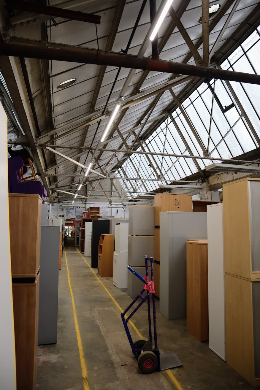 An alternative view along ORS's warehouse roof structure showing the newly installed linear LED lighting and roof trusses above rows of stacked office furniture.