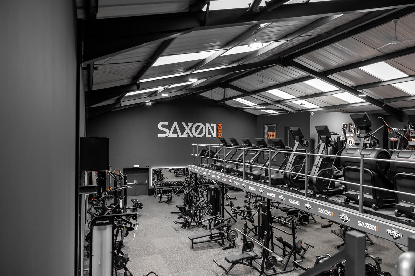 Raised cardio deck at Saxon Gym in Cannock, with a row of treadmills and cross-trainers served by dado trunking along the platform edge.