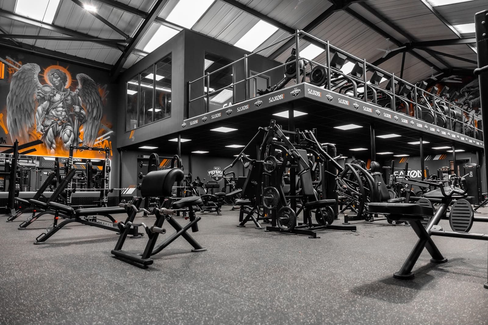 Main gym floor at Saxon Gym in Cannock, showing LED panel lighting across the suspended ceiling beneath a mezzanine level, with resistance machines, free weights and a painted mural on the far wall.