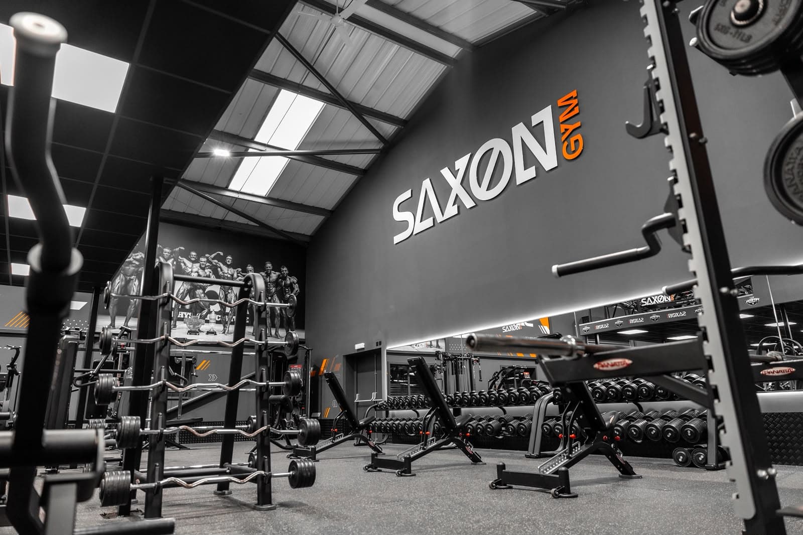 Free weights area at Saxon Gym in Cannock, with dumbbell racks along a mirrored wall lit by a continuous linear accent light, beneath rooflights and an exposed steel roof structure.