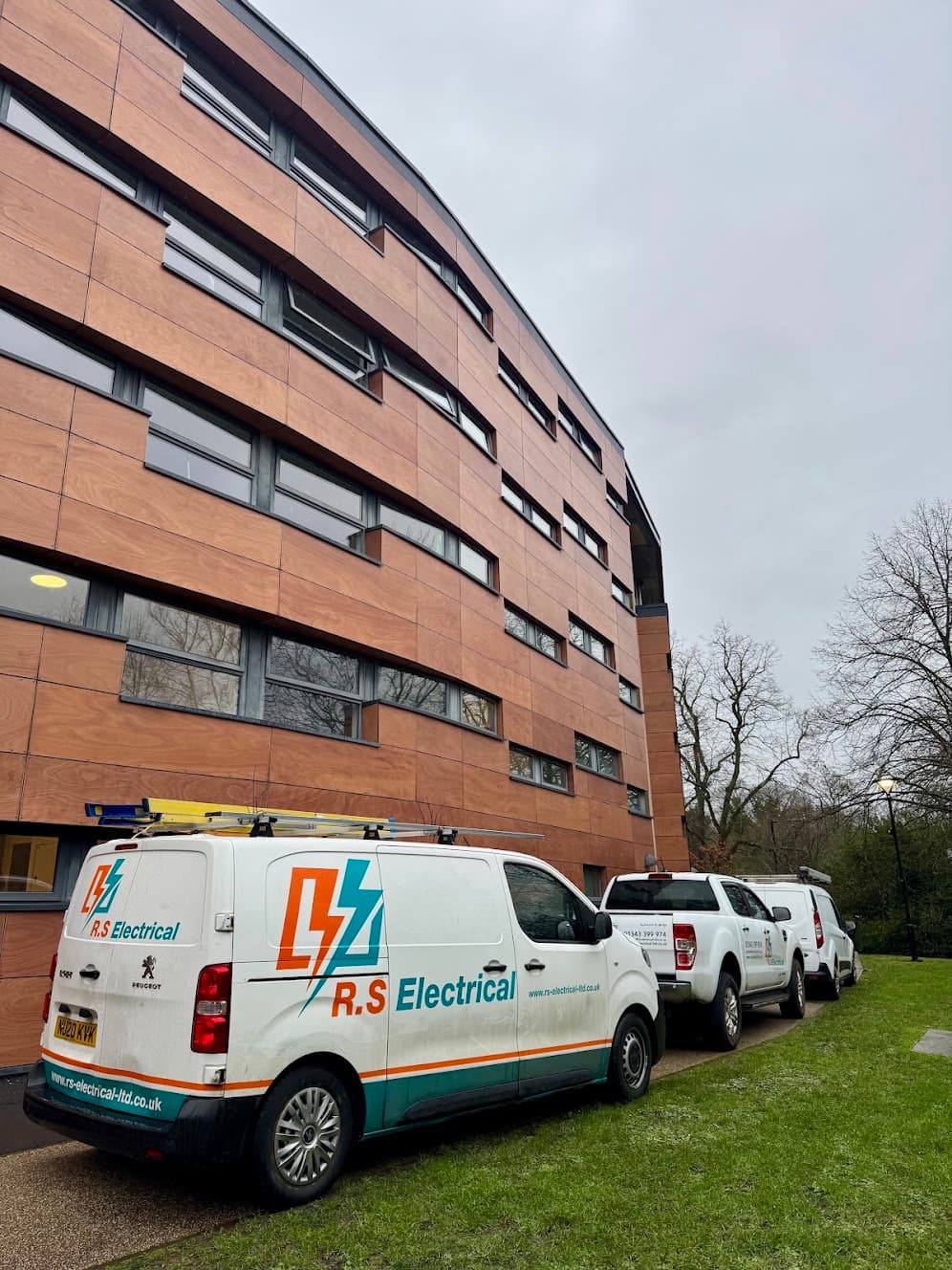 Three RS Electrical liveried vehicles parked outside a modern University of Birmingham building with timber-clad exterior panelling.