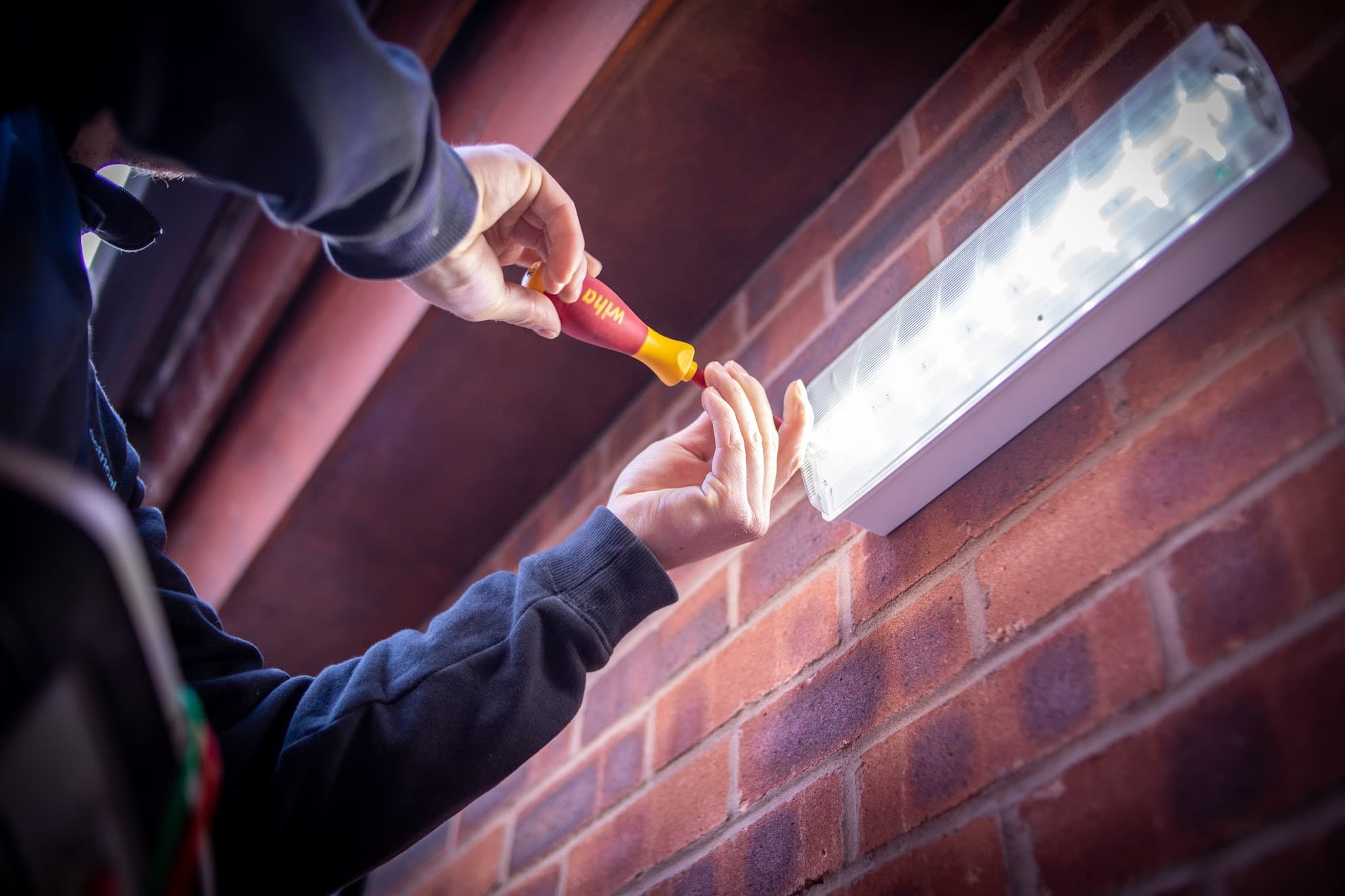 An RS Electrical engineer using an insulated screwdriver to service a wall-mounted emergency light fitting on an exposed brick wall.